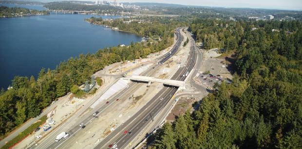 An aerial shot of a major roadway running through a forested area with a body of water on the left.