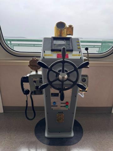 Ship's helm with a wheel, brass periscope, and controls in front of a window overlooking the sea.