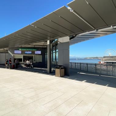 Photo shows Washington State Ferries customers lined up at the ticket booth at Colman Dock in Seattle on a sunny day. In the background, there is Puget Sound and the Ferris Wheel. 