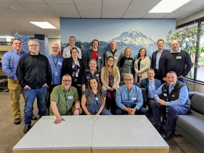 19 people posed in an office setting with a mountain mural in the background