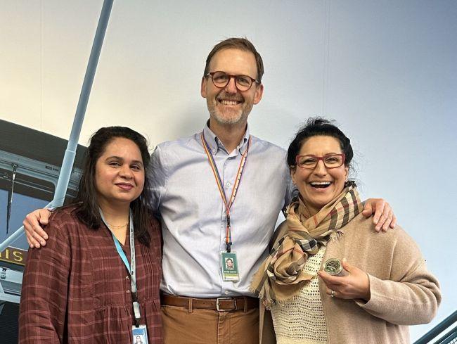 Three people smiling and standing together in front of a light blue background with one holding up a coin