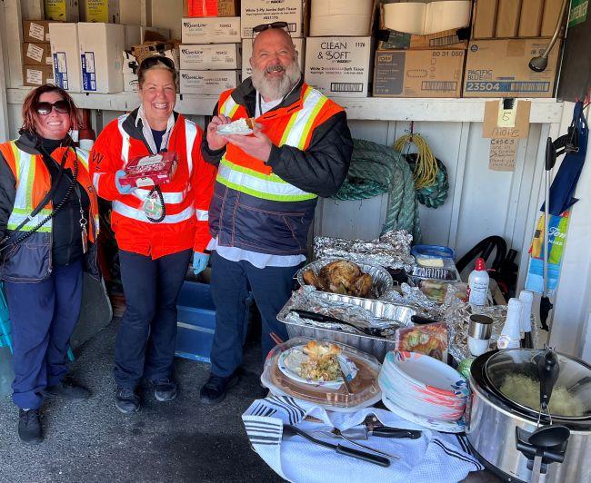 Three people in safety vests standing in a storage room with a table of food in front of them.