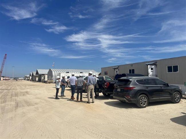A group of people stands beside two parked vehicles in an open sandy area, with prefabricated buildings and a crane visible in the background under a blue sky.