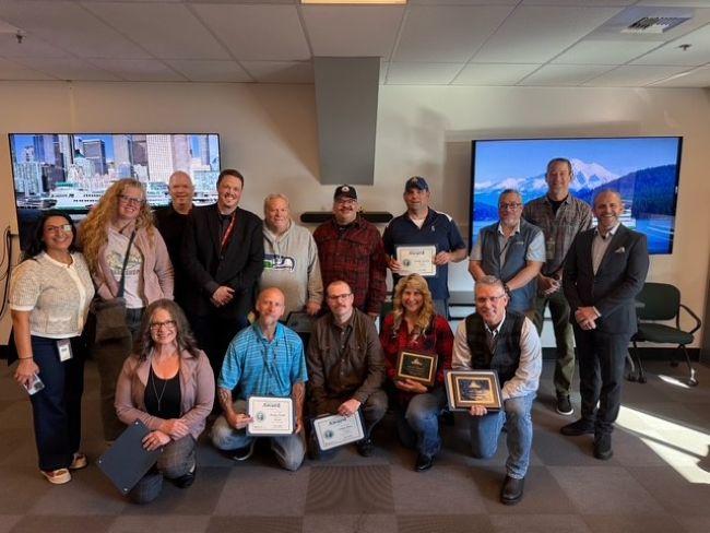 A group of people in a room holding certificates and plaques with two monitors displaying a city skyline and mountain landscape.